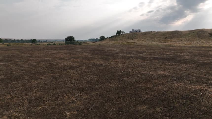 A wide dry field stretches toward a low hill under a cloudy sky