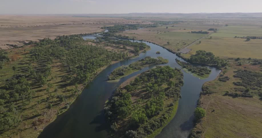 An aerial view captures a wide river winding through a flat landscape with scattered trees lush fields and dry undulating hills in the distance under a bright sky