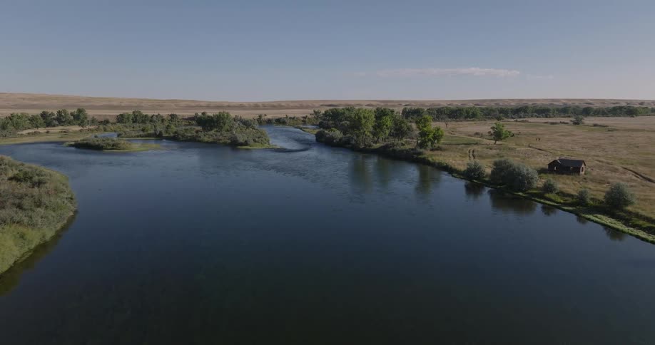 An aerial view showcases a calm river or lake winding through expansive fields and trees with a small cabin visible on the far bank