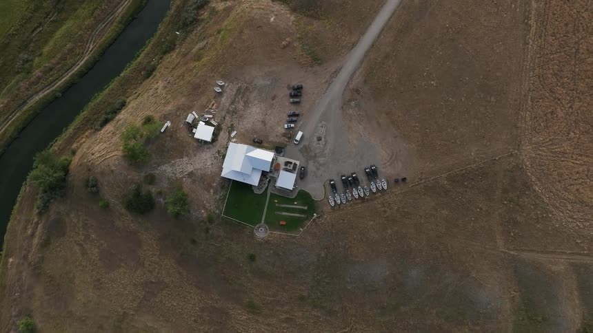 An aerial view shows a large building with a white roof on a hill next to a narrow canal surrounded by dry land and parked vehicles