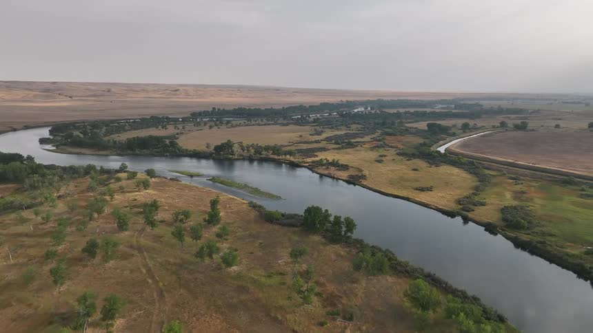 An aerial view follows a wide river as it winds through a vast dry valley with trees clustered along its banks under a hazy sky
