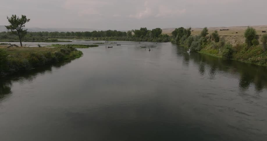 People are fishing in boats on a calm river surrounded by trees and open fields
