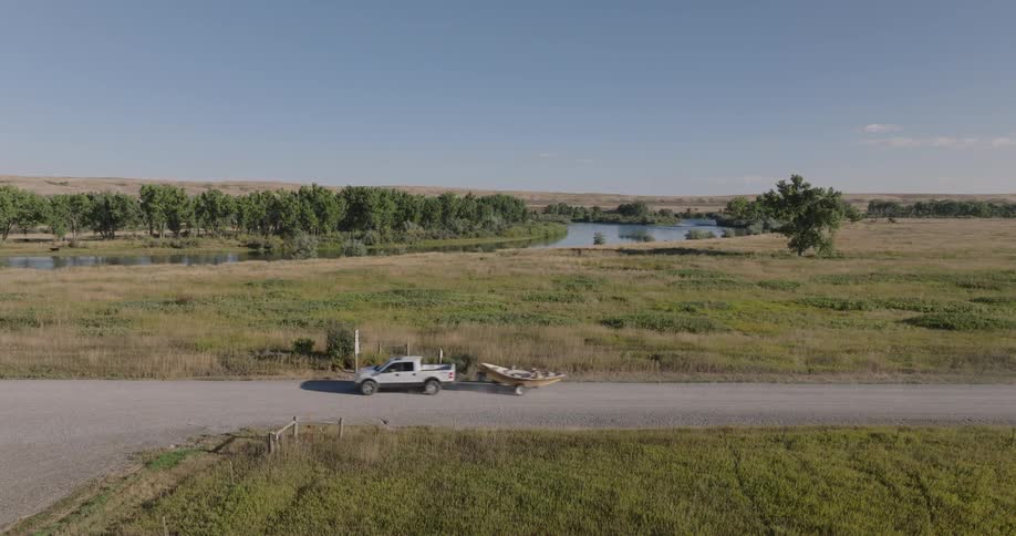 A white truck hauls a small boat along a rural road beside a grassy field and a calm river