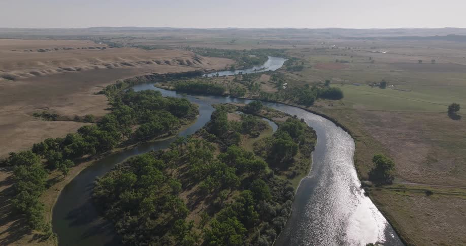 An expansive aerial view captures a winding river flowing through a lush green landscape with surrounding hills under a bright sky