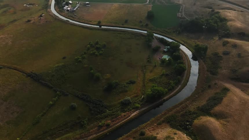 An aerial view shows a narrow river winding through a rural landscape with fields and scattered buildings under a cloudy sky with visible sunrays