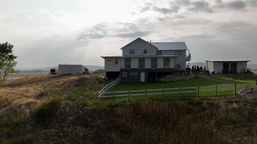 A rural farmhouse sits on open land with a fire pit fenced yard and small outbuilding nearby