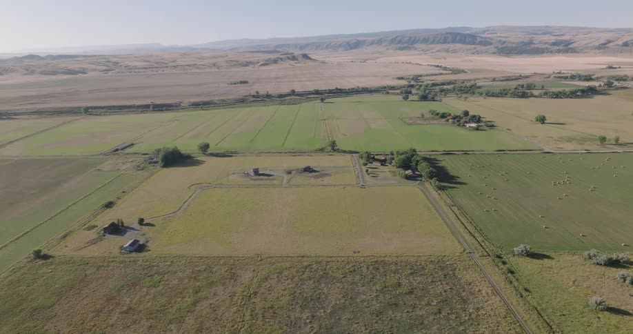 An aerial view showcases a vast landscape of patchwork fields some with hay bales and scattered farm buildings under a clear sky with distant mountains