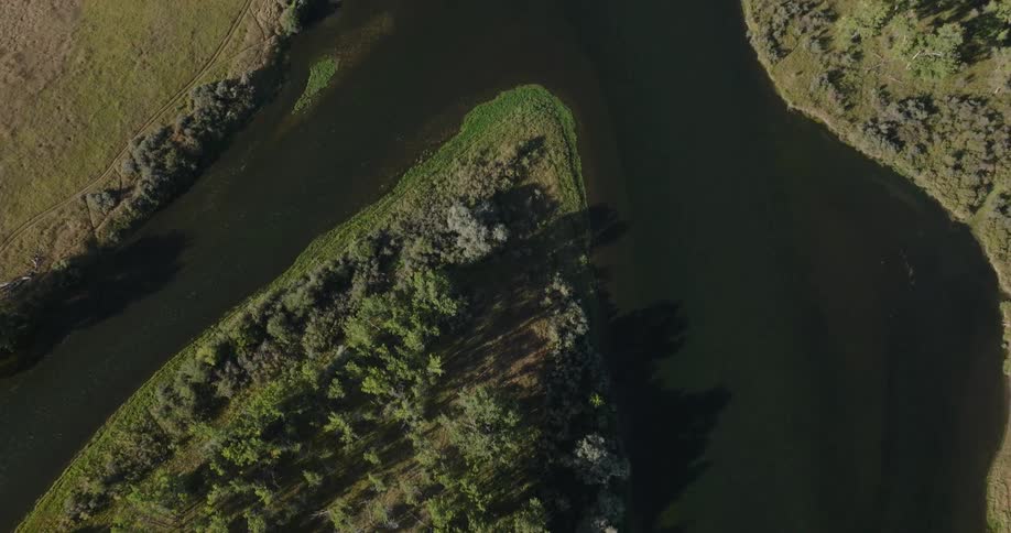 An aerial view looks down on a dark river as it curves through a landscape of trees and grassy hills casting long shadows in the sunlight