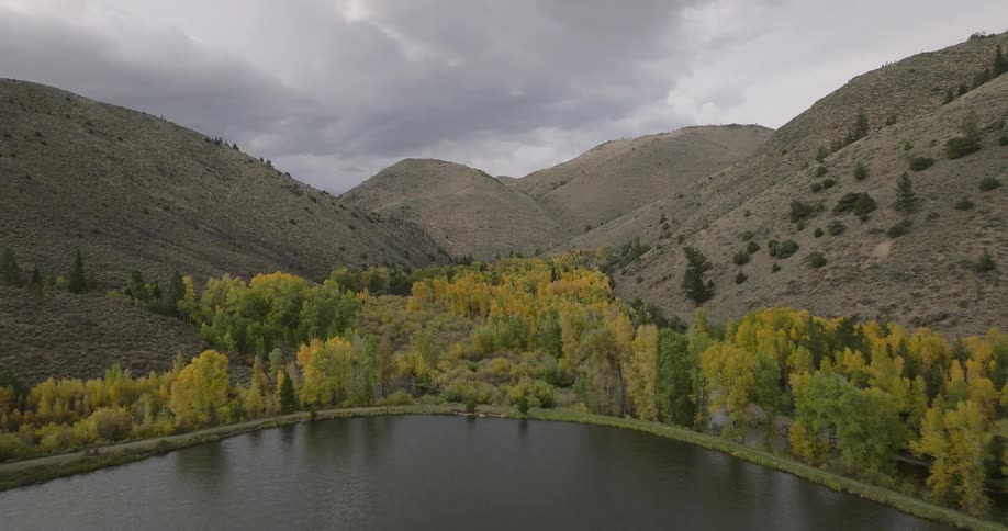 A calm lake is surrounded by colorful autumn trees and dry mountains under a cloudy sky