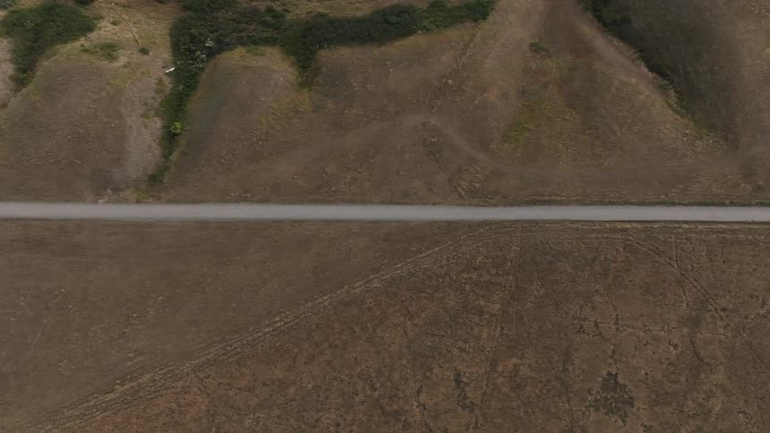 A topdown aerial view shows a paved road cutting through a dry brown hilly landscape with sparse green vegetation