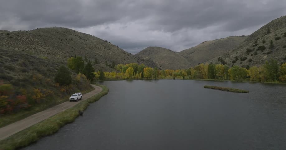 A white car drives along a lakeside road surrounded by hills under a cloudy sky
