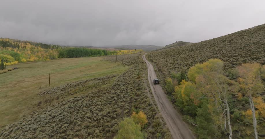 A car drives down a dirt road through a remote landscape with hills trees and cloudy skies