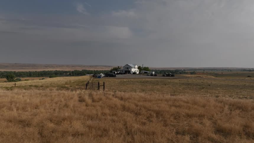 A white farmhouse sits isolated in a vast dry field under a cloudy sky