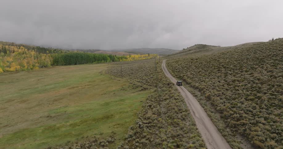 A vehicle drives down a dirt road through a vast hilly landscape under an overcast sky