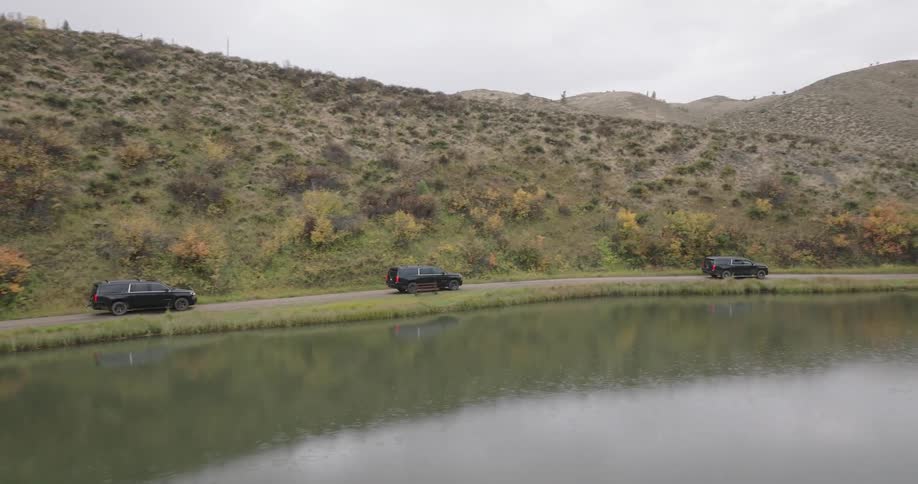 Cars drive beside a pond near autumn trees and boats with hills in the background