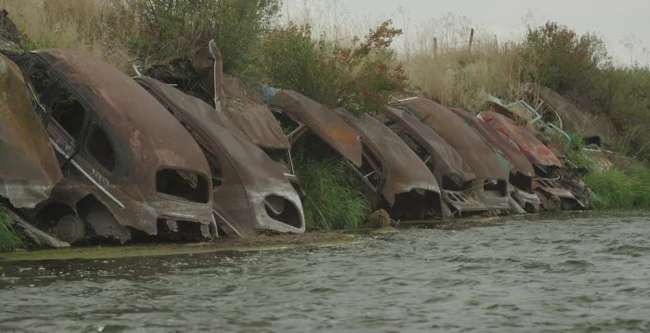 A row of rusted abandoned car shells lines the edge of a riverbank