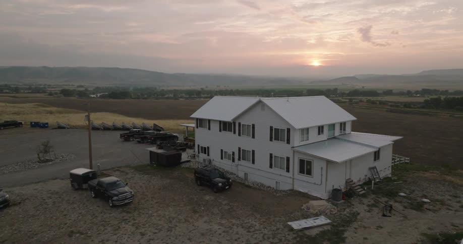Vehicles parked around a white house in a field area with the sun on the horizon