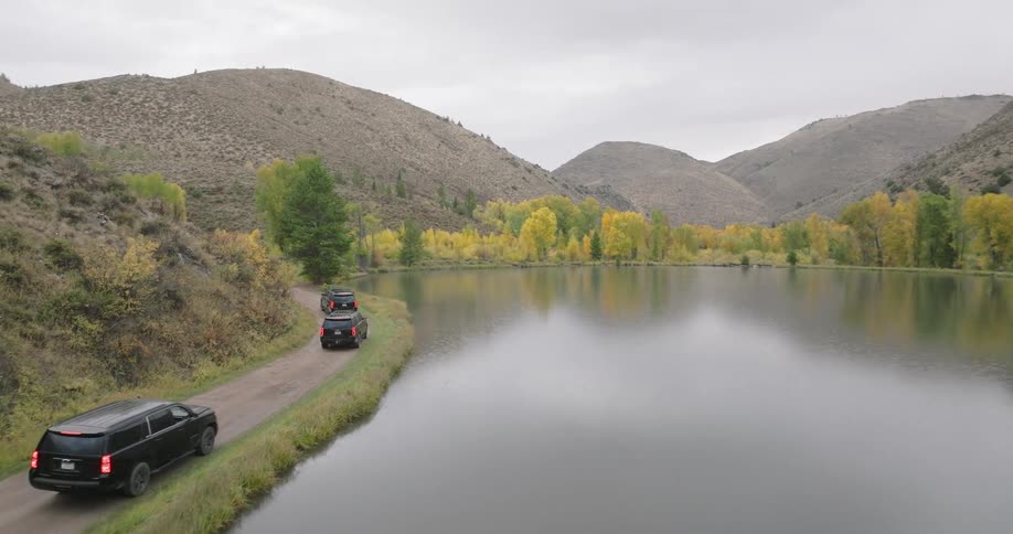 Cars drive along a dirt road beside a calm lake surrounded by autumn trees and hills