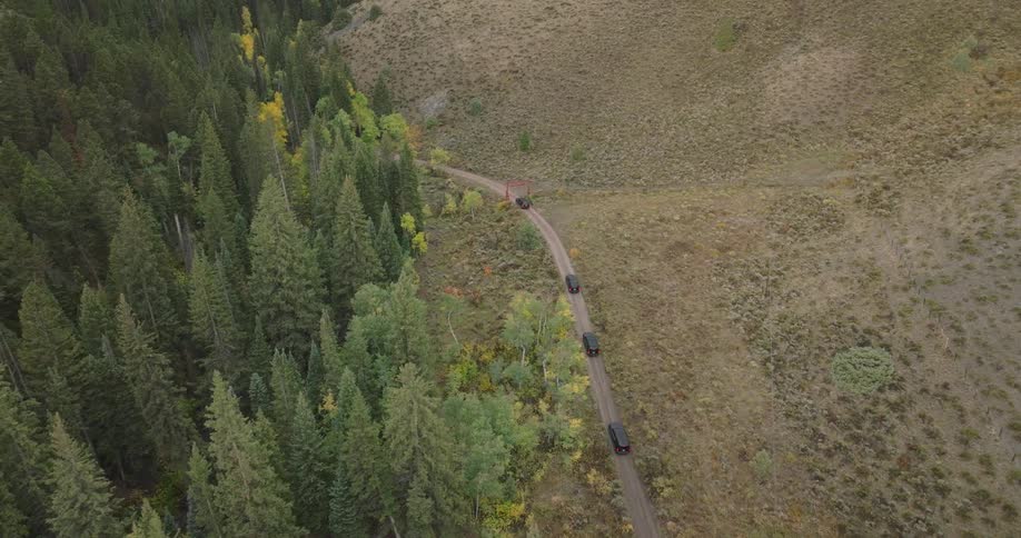 An aerial view flying lower to the road reveals multiple black vehicles on a rural dirt road that goes under a red gate surrounded by a mountainous forest with hints of autumn colors