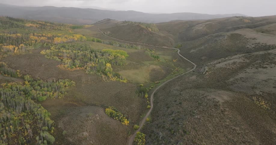 An aerial view captures a winding road through a valley flanked by hills and patches of trees displaying autumn colors