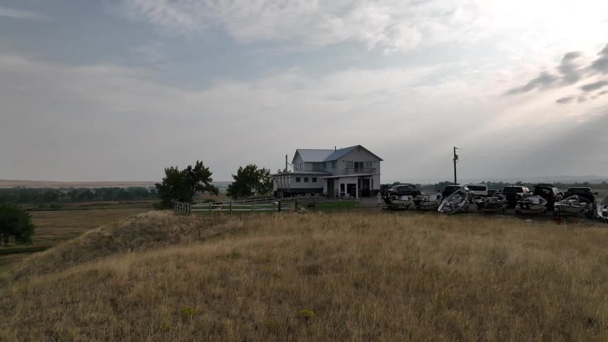 A house sits on a grassy hilltop with several parked vehicles beside it under a cloudy sky