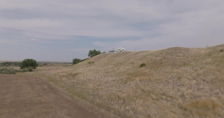 A white house with a wraparound porch sits atop a dry grassy hill under a cloudy sky