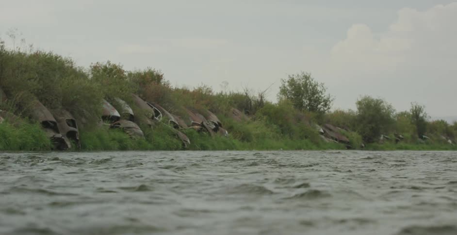 A calm river flows past an overgrown bank with partially buried concrete structures