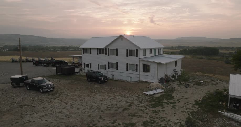 A white farmhouse sits in a rural landscape at sunrise with trucks and boats nearby