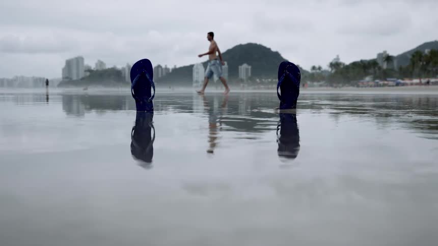 Two dark blue flipflops stand upright on a wet sandy beach as a man walks in the background with distant buildings and mountains beneath an overcast sky