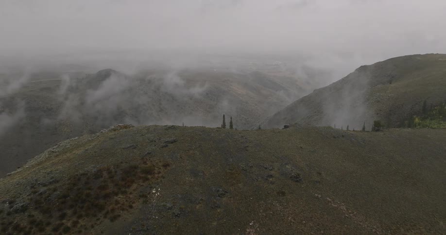 Fog rolls through a rugged mountain valley covered in sparse vegetation and trees