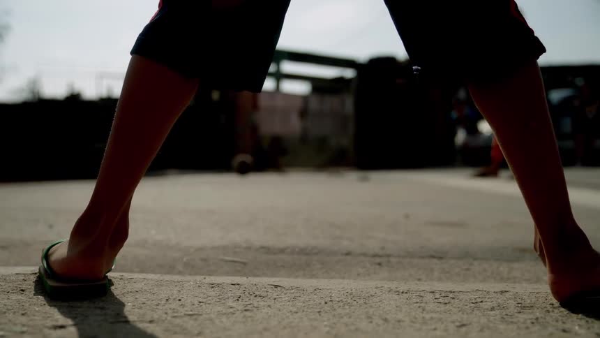 Child wearing flipflops with legs open in the foreground and in the background a person ready to kick a ball