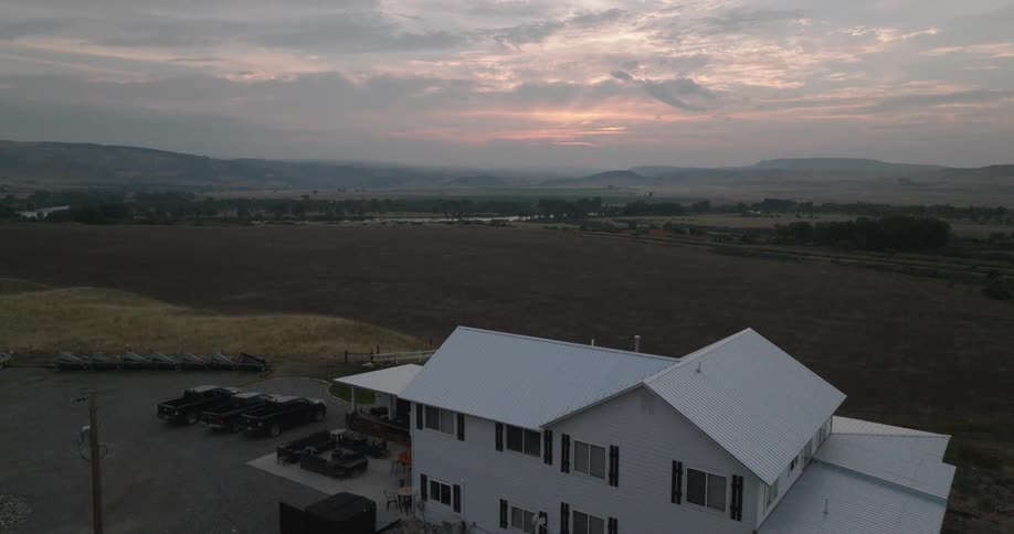 A large white farmhouse sits beside open farmland under a cloudy sunset sky