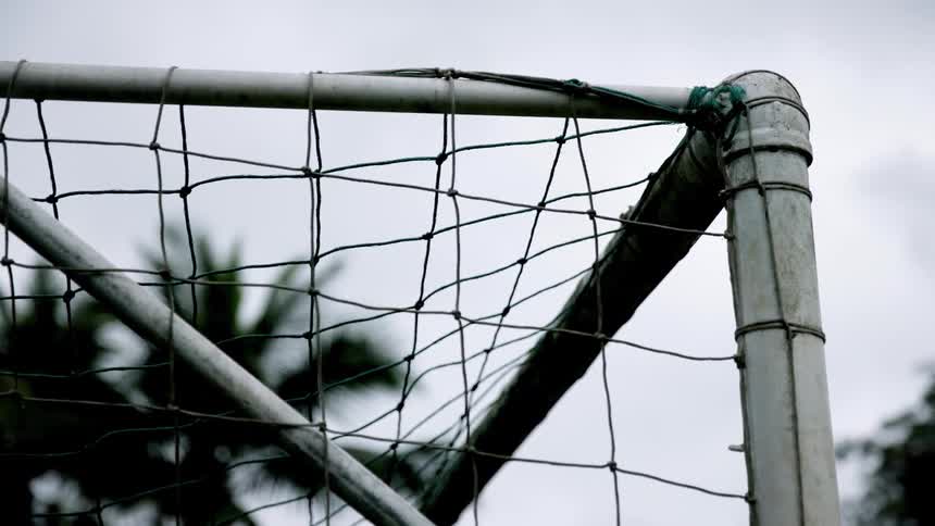 Up close on the worn top corner of a soccer goal post the net hangs in front of an overcast sky and a distant palm tree