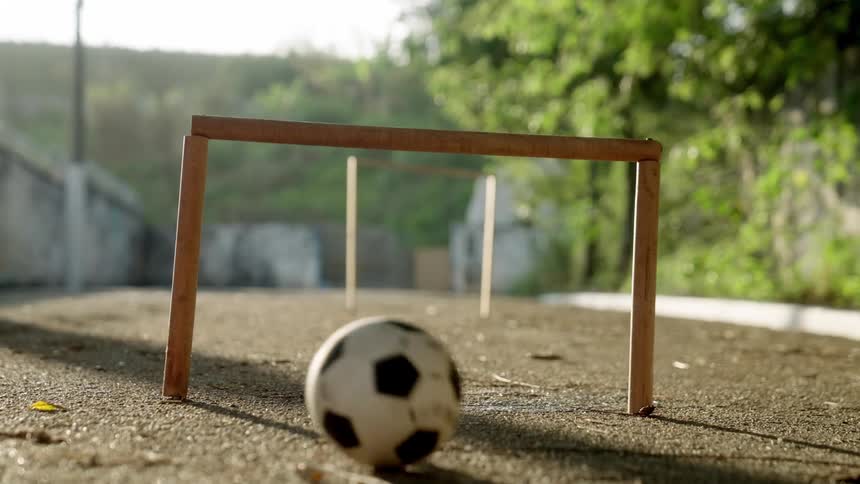 A soccer ball rolls past a small wooden makeshift goalpost positioned on a road during daytime