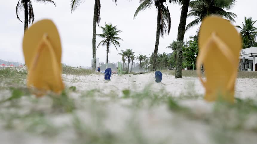 Yellow slippers are stuck in the beach sand surrounded by grass with another pair of blue slippers in front palm trees line the area under the midday sun
