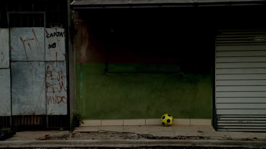A yellow soccer ball sits alone in front of a weathered green wall in an urban street setting