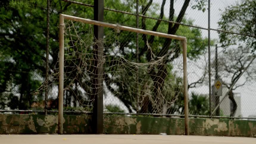 An abandoned soccer goal with a torn net stands on a paved surface with trees and a fence in the background