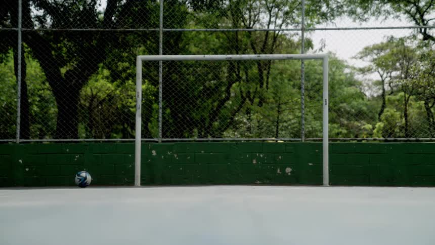 A soccer ball rolls on the ground next to an empty goal on an outdoor court