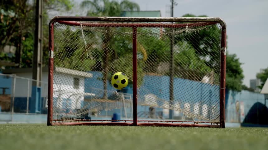 A ball is played into a small netted goalpost on an outdoor soccer field during daytime