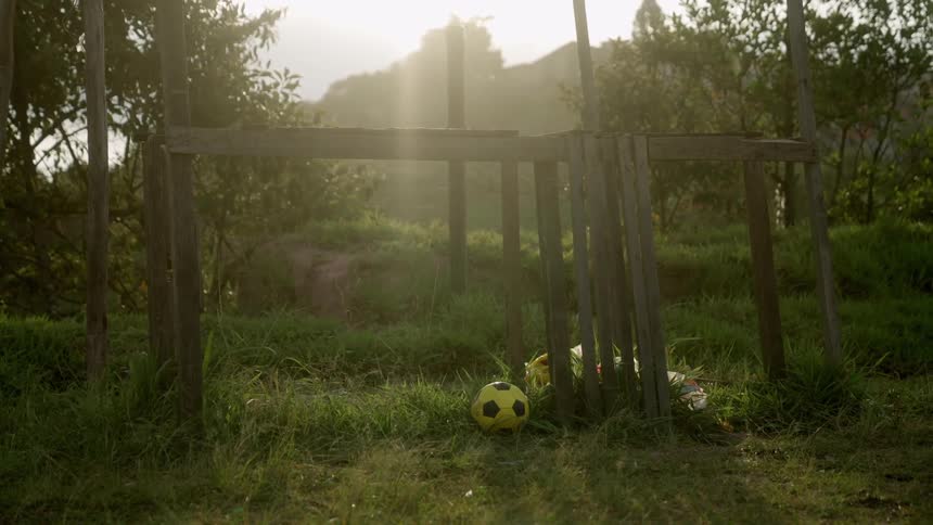 A yellow soccer ball sits in the tall grass before a handmade wooden goal as bright sunlight streams through the background