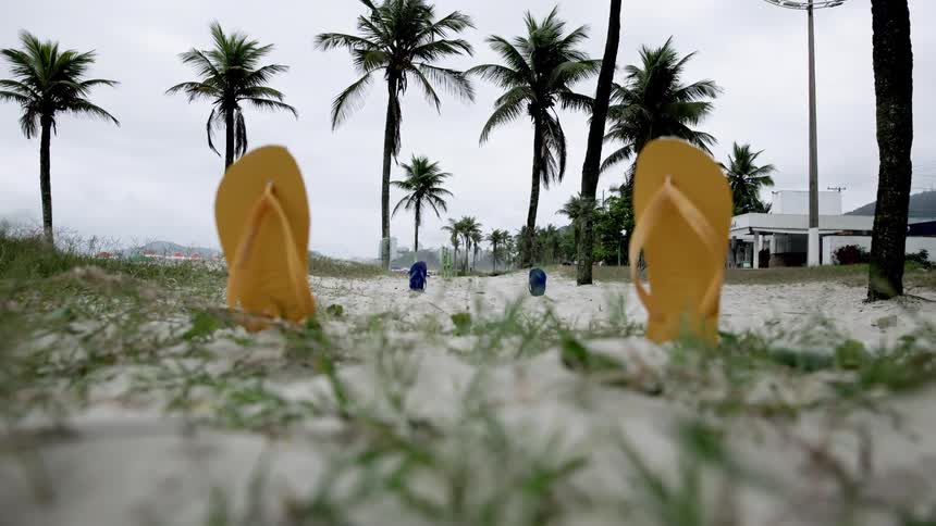 Two yellow flip flops stand upright in the sand on a beach with palm trees and a cloudy sky in the background