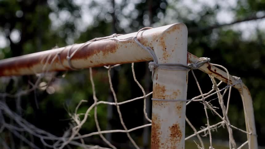 A closeup shot shows the corner of a rusted and weathered soccer goal post with its tattered net tied on with wire