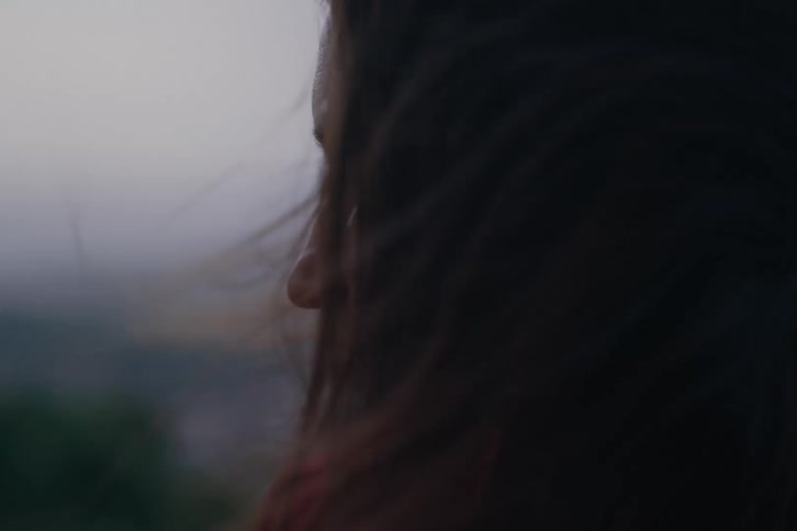 Woman with long hair stands in profile before a hazy hillscape her hair lifted gently by the wind in warm low light