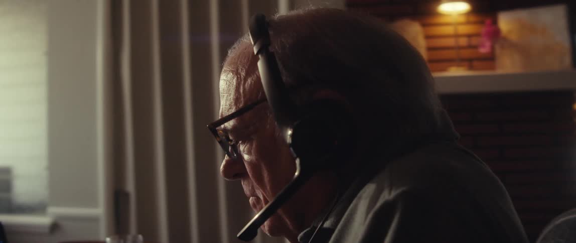 Elderly man wearing headphones works on a laptop seated in a dim room lit by warm hanging lamps