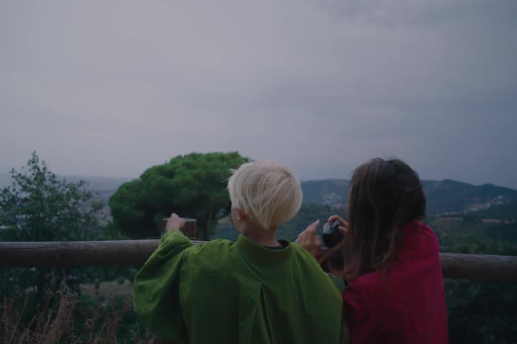 Two children stand by a wooden railing as one points toward distant mountains beneath an overcast sky
