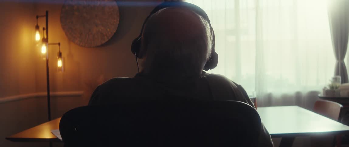 Balding man leans back in his chair while sitting at a table and listening to headphones