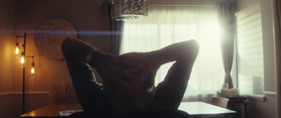 A person sits at a dining table with hands clasped behind the head facing sunlit sheer curtains under a chandelier