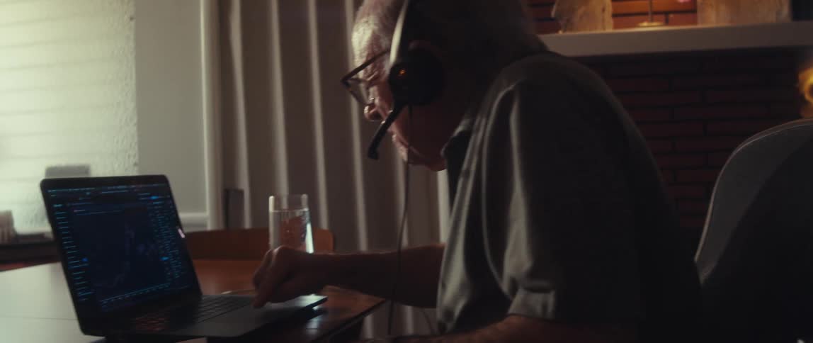 An older man wearing headphones and glasses intently looks at a laptop screen possibly working or engaged in a video call