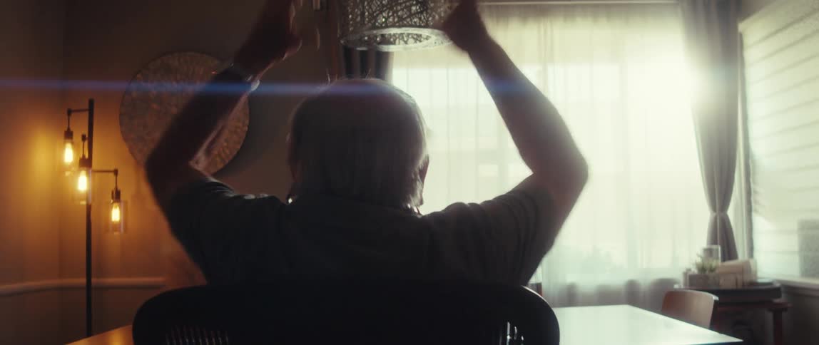 An elderly man raises his arms in joy while sitting at a desk in a sunlit room