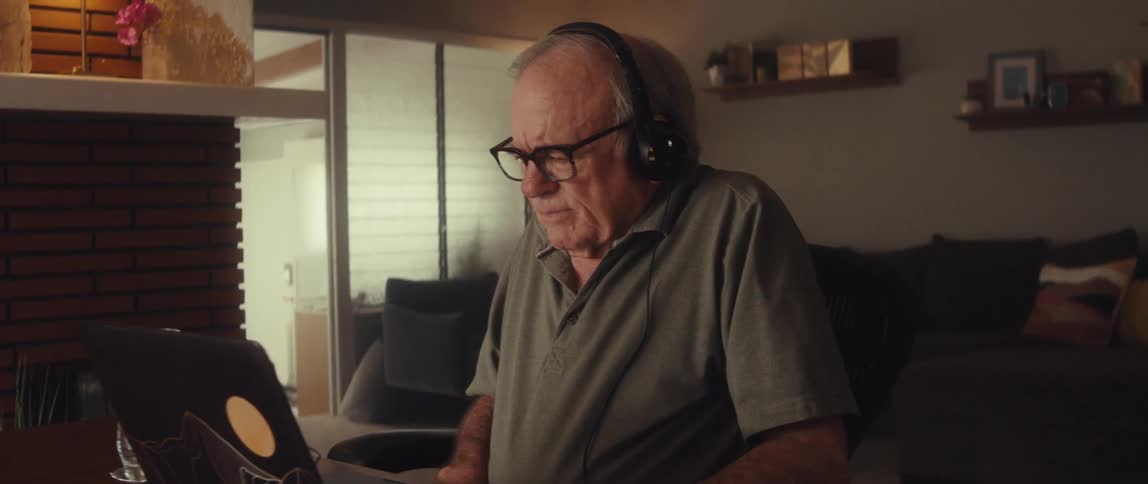 An elderly man wearing headphones focuses on his laptop during a video call at home